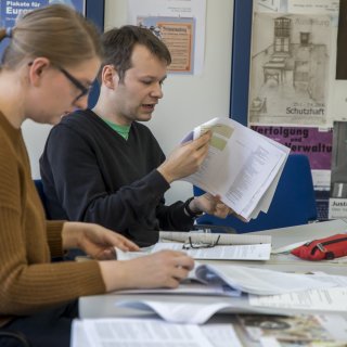 Eine Frau und ein Mann lesen in Zeitungen und Zeitschriften, die sie vor sich auf dem Tisch ausgebreitet haben. An der Wand hinter ihnen hängen verschiedene Plakate.