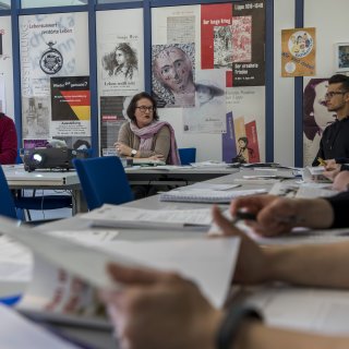 Archivpädagog*innen unterhalten sich mit Besucher*innen in einem Konferenzraum. Dieser ist mit bunten Postern zu geschichtlichen Ausstellungen geschmückt.