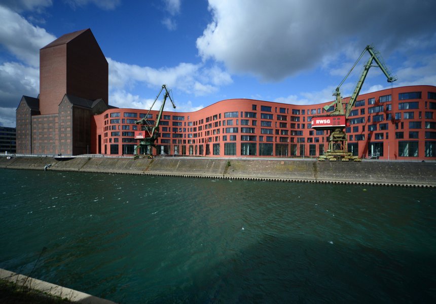 Außenansicht des Dienstgebäudes in Duisburg mit dem Magazinturm und dem wellenförmigen Verwaltungsgebäude, davor zwei Kräne und der Kanal des Innenhafens. 