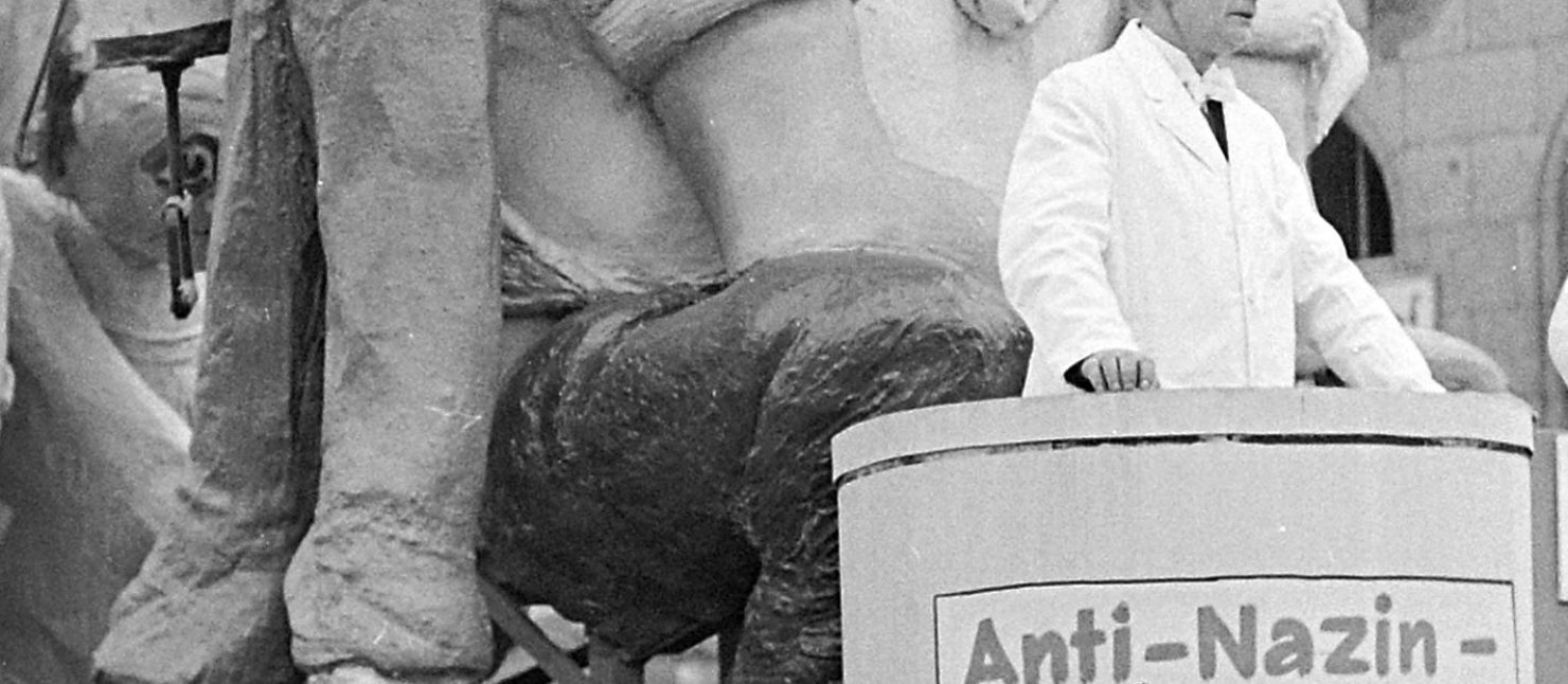 Mottowagen "Tünnes wird entnazisiert auf dem Kölner Rosenmontagszug 1950 Titelbild der Ausstellung. Zu sehen ist ein Karnevalswagen auf dem "entnazisiert" steht. Ein Patient bekommt eine Spritze.