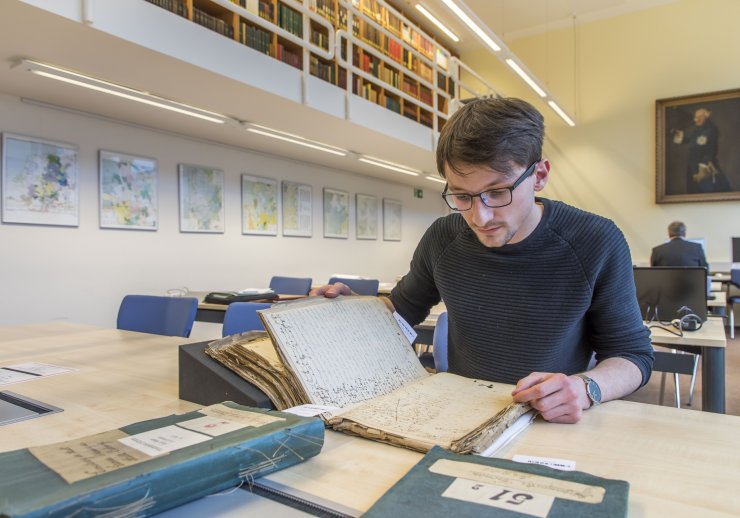 Ein Benutzer sitzt im Lesesaal. Vor sich hat er ein sehr altes und fragiles Buch aufgeschlagen. Um das Buch zu schonen, wird es durch einen speziellen Keil gestützt.